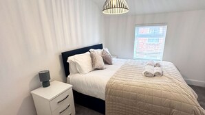A neatly made bed with a quilted cover, a bedside lamp, and a window with blinds. - The Little Tern - A Cosy Coastal Retreat (Lancashire)