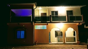 A two-story building with a balcony on the upper floor, a small entrance with a door, and a window with blue curtains. - Serenity Villa in Sunny Burusbi Phase 2 (Sukuta)