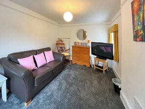 A living room with a dark leather sofa, a wooden dresser, a television on a stand, a round mirror, and a painting on the wall.