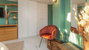 Ein modernes Schlafzimmer mit türkisfarbenen Wänden, einer Holzkommode, einem roten Samtstuhl und einem großen Spiegel, der den Raum reflektiert.