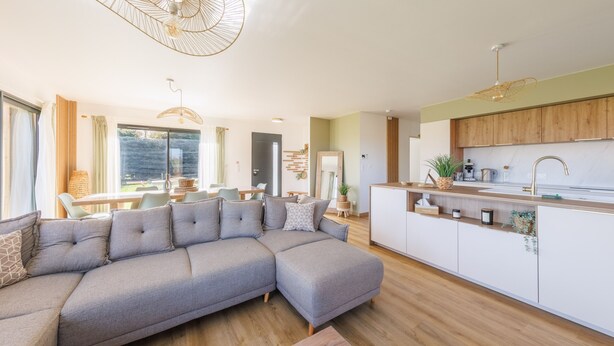 Ein modernes Wohnzimmer mit einem grauen Ecksofa, Holzboden und einer Küche mit weißen Schränken und einer Holz Arbeitsplatte.