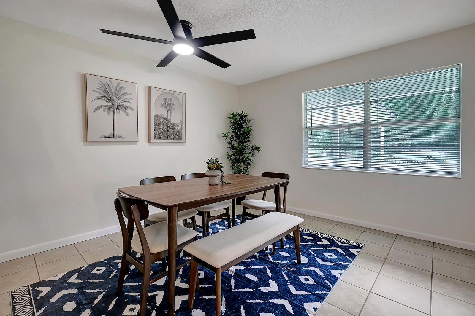 A dining area with a wooden table, chairs, a bench, a ceiling fan, and a window with blinds.