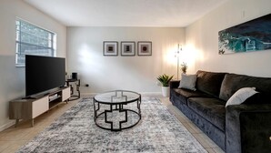 A living room with a flat-screen TV, a dark-colored sofa, a glass coffee table, and a large painting on the wall.