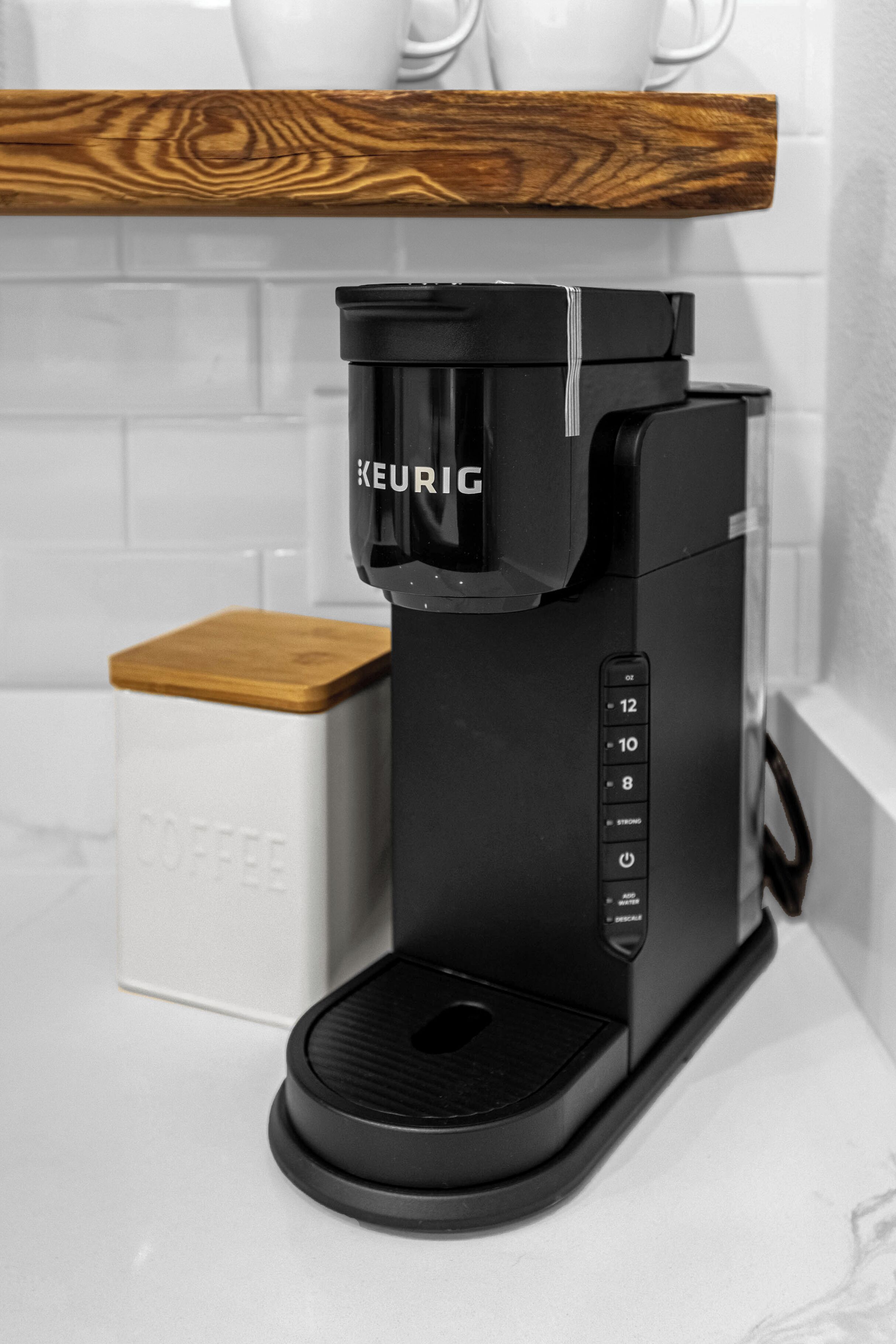 A black Keurig coffee maker on a white countertop with a wooden shelf above it.