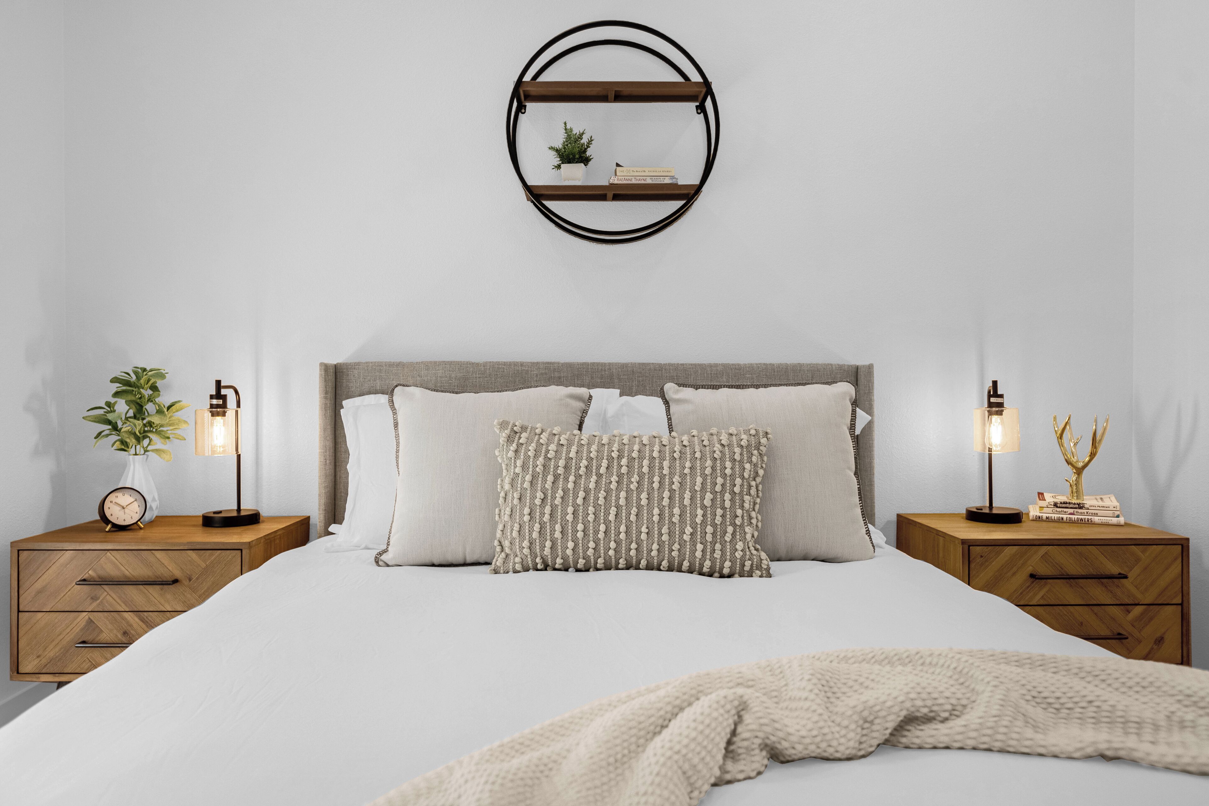 Un lit bien fait, recouvert d’une couette blanche et d’un plaid tricoté. Deux tables de chevet, sur lesquelles sont posées des lampes, se trouvent de chaque côté du lit, et une petite horloge est posée sur l’une d’elles. Un miroir rond est accroché au-dessus du lit.