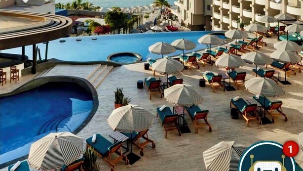 Zona de piscina en un resort con múltiples tumbonas y sombrillas, una gran piscina y vistas al mar y a las montañas de fondo.