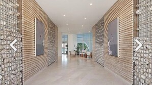 Un pasillo de diseño moderno, con una pared decorada con un motivo en un lado y una pared lisa en el otro. En la pared lisa hay obras de arte enmarcadas, y el pasillo conduce a una zona de estar con mesas y sillas.