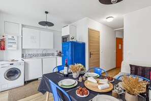 A compact kitchen with a washing machine, a blue refrigerator, and a dining area with a table set for two. - Lavie Maison Cozy 3BR Haven w Parking (Bordeaux)