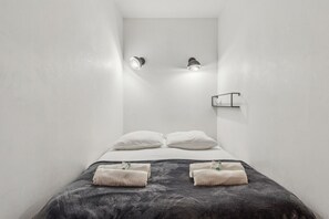 Une chambre d’hôtel minimaliste, avec un lit, deux lampes fixées au mur, une petite étagère et deux serviettes posées sur le lit.