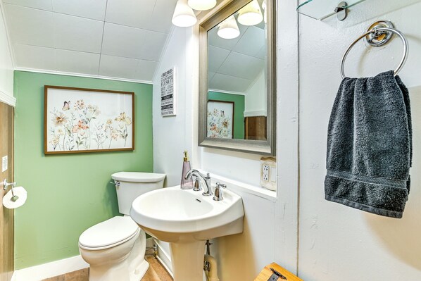 Un baño con inodoro, lavamanos y espejo. Se observan cuadros con motivos florales en la pared y una toalla colgada de un gancho.