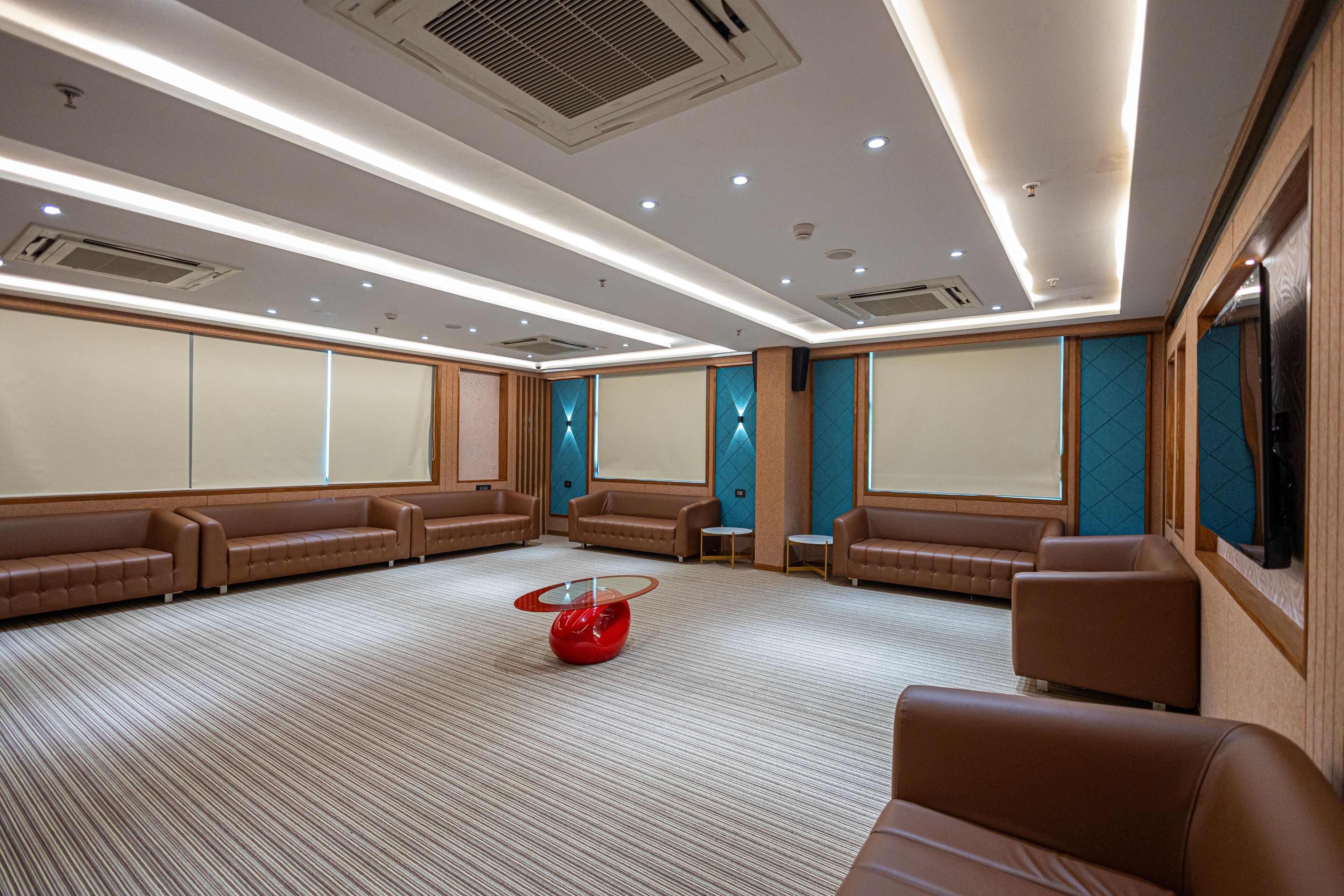 A spacious room with multiple leather sofas arranged around a central red table, recessed lighting, and a large flat-screen TV mounted on the wall.