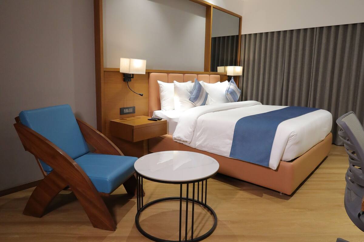 A hotel room with a bed, a blue chair, a small table, and a nightstand with a lamp.