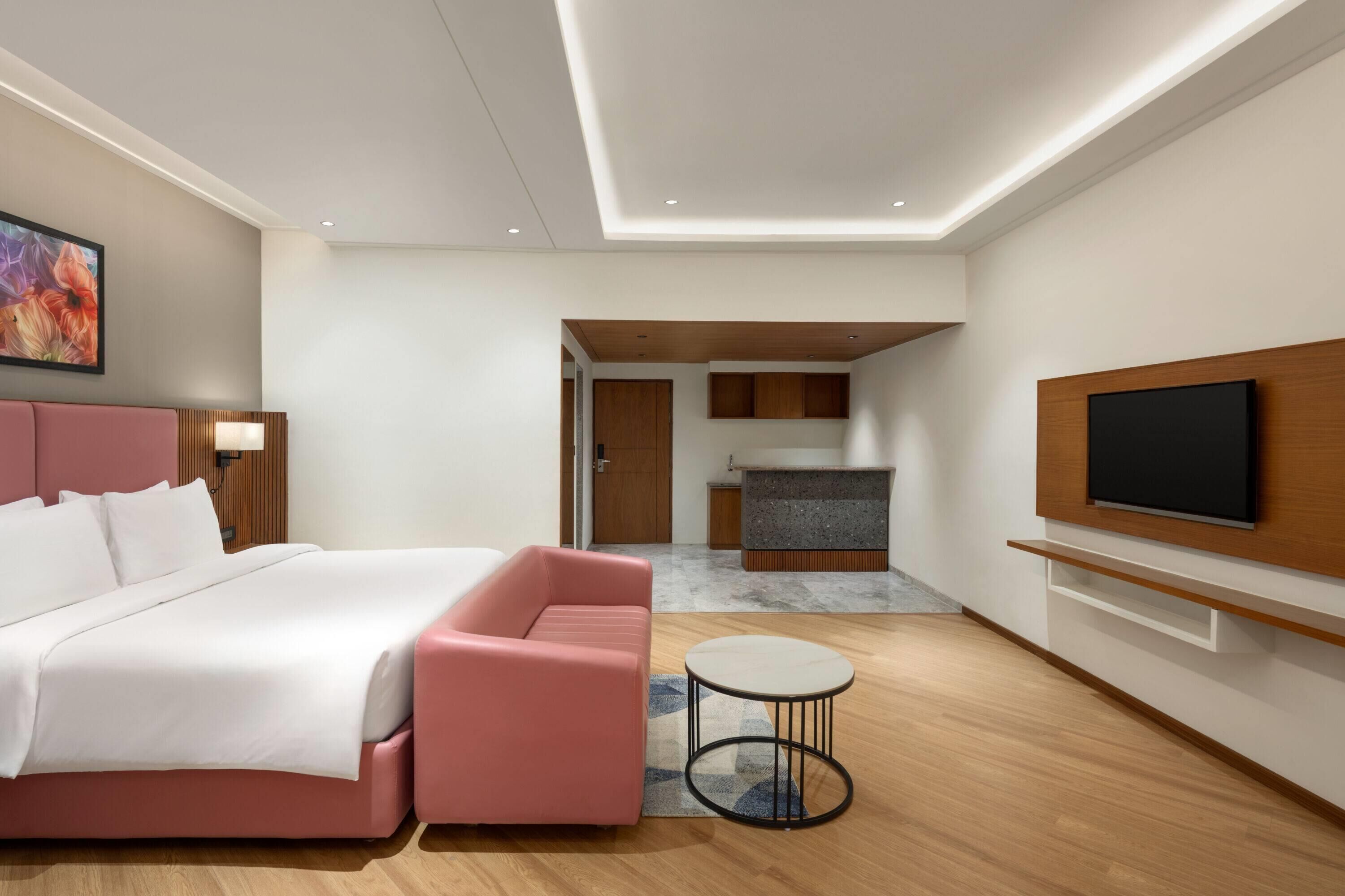 Una habitación de hotel moderna con una cama, un sofá rosa, una pequeña mesa redonda, un televisor montado en la pared y una zona de cocina al fondo.