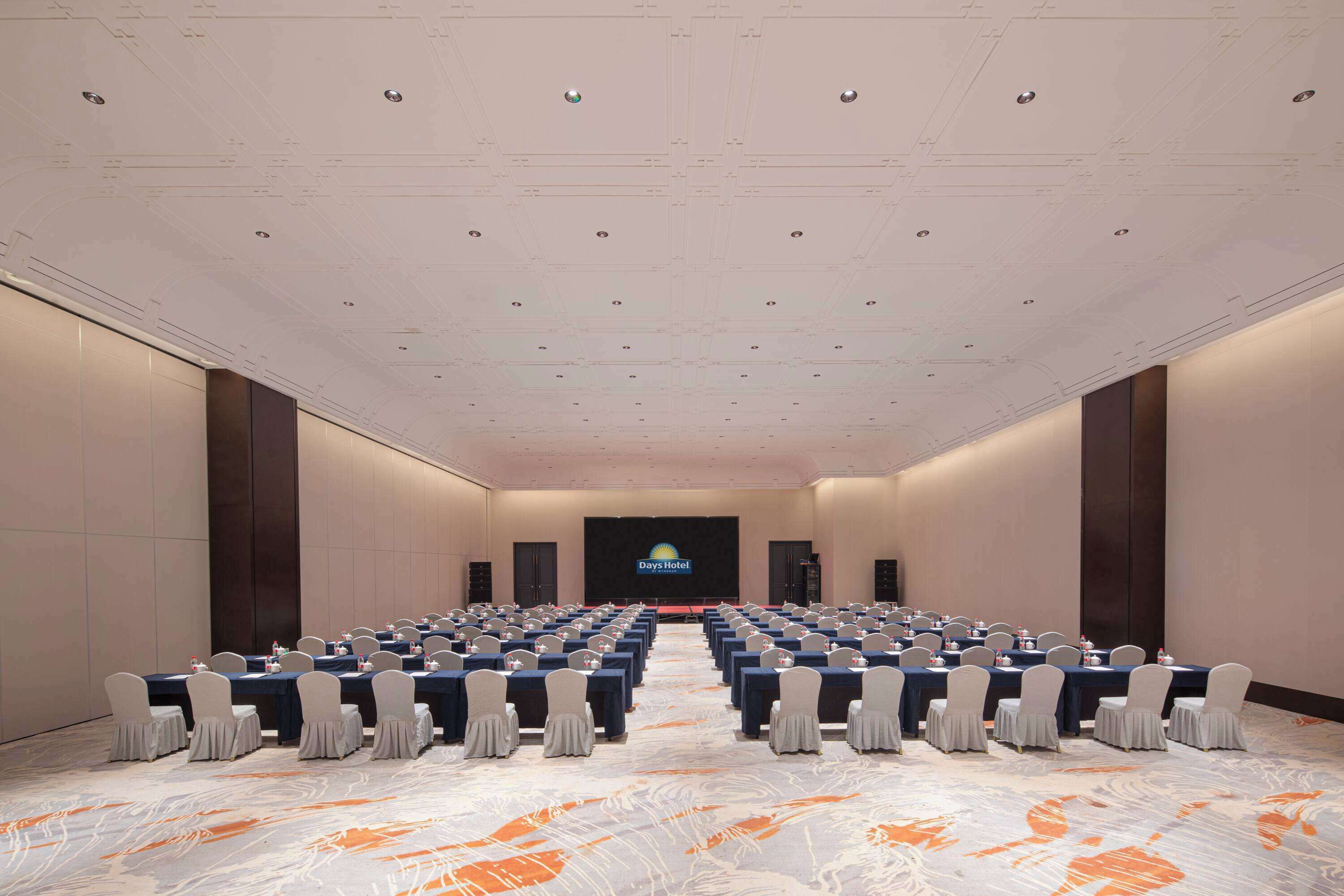 A conference room with rows of tables and chairs, a large screen at the front, and a patterned carpet.