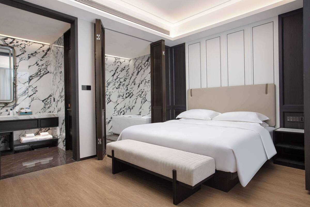 A modern hotel room with a marble bathroom, a large bed with a headboard, and a bench.