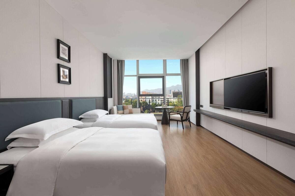 A hotel room with two beds, a TV, a small table, and a window with a city view.