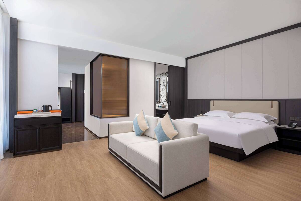 A modern hotel room with a sofa, a bed, a nightstand, a desk with a kettle, and a large mirror.