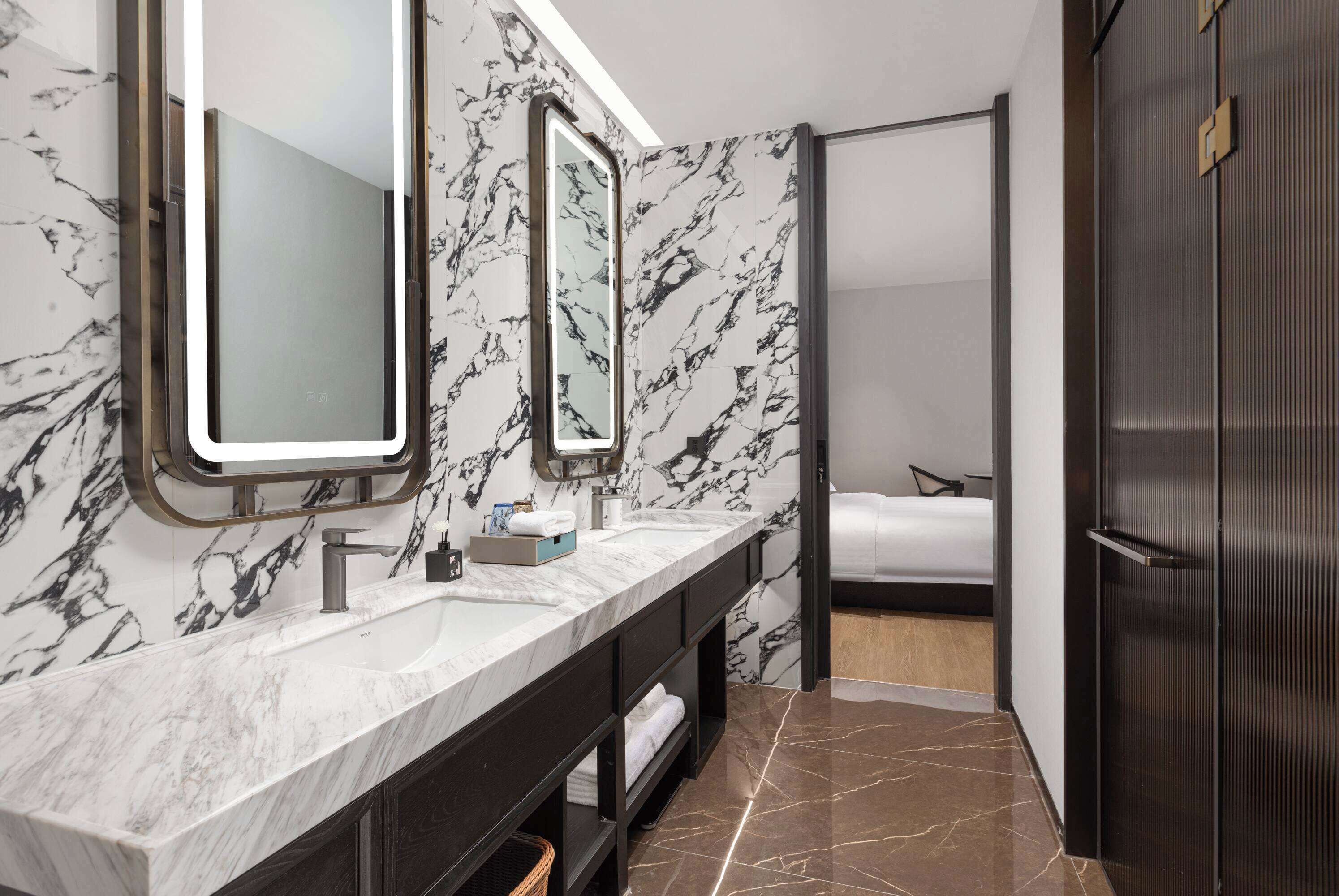 A modern bathroom with marble walls, a large mirror, a double sink, and a bed in the adjacent room.
