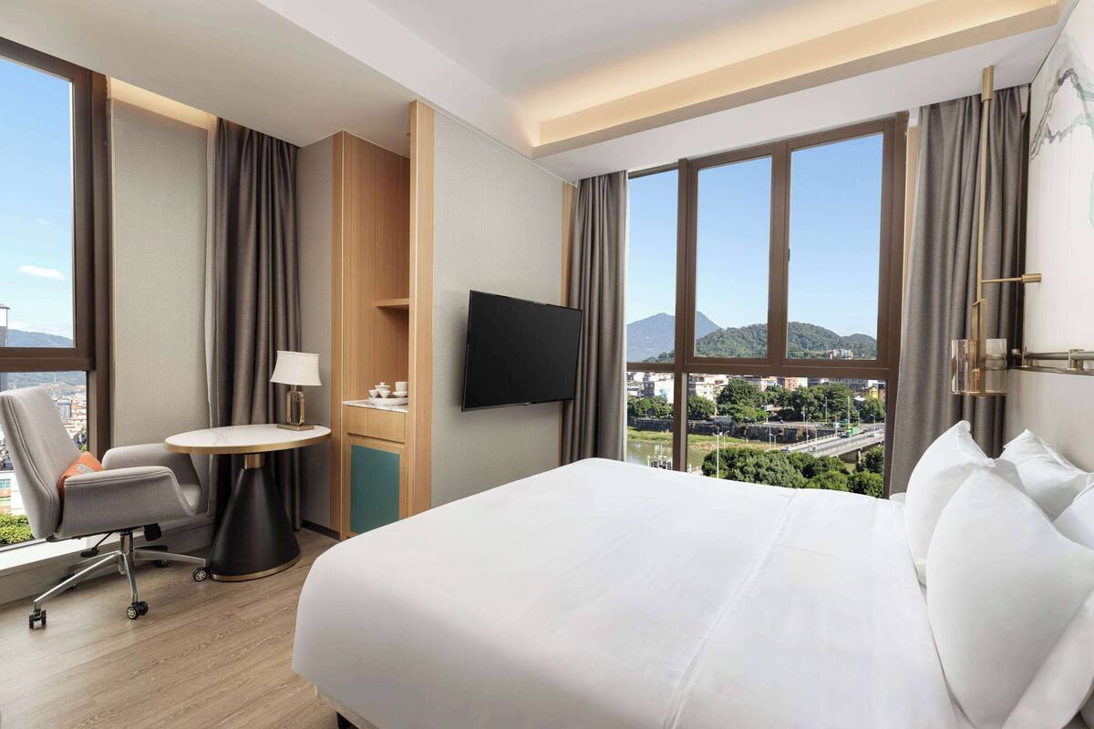 A hotel room with a large bed, a desk with a lamp, a TV, and a view of a cityscape through large windows.