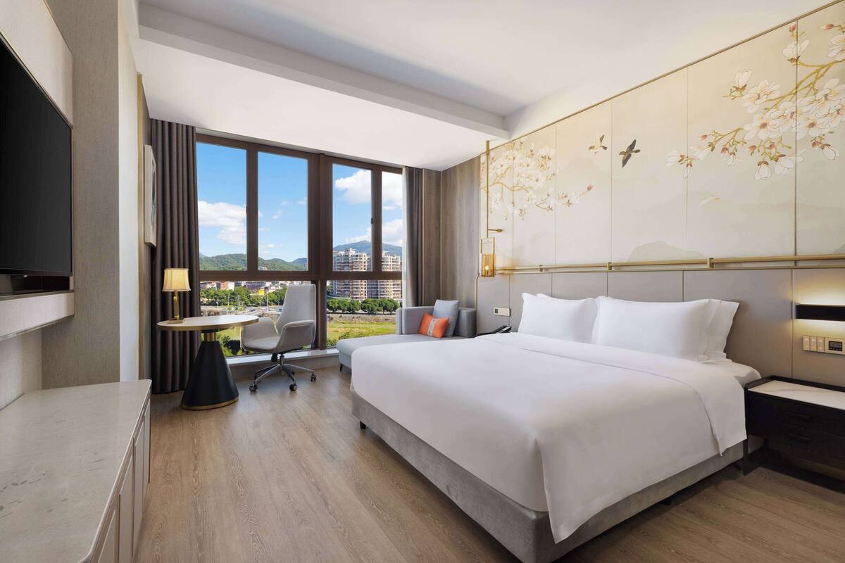 A modern hotel room with a large bed, a desk with a chair, a TV, and a window with a city view.