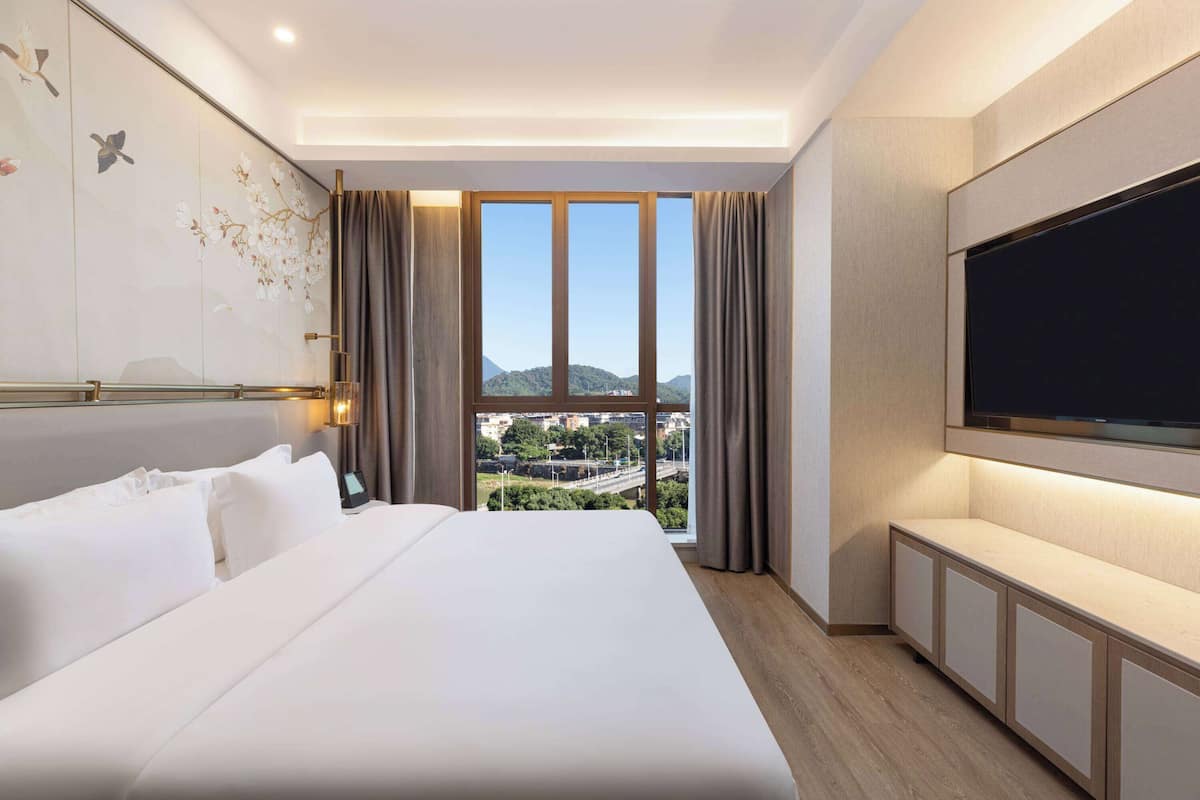 A hotel room with a large bed, a TV, and a view of a cityscape through a large window.