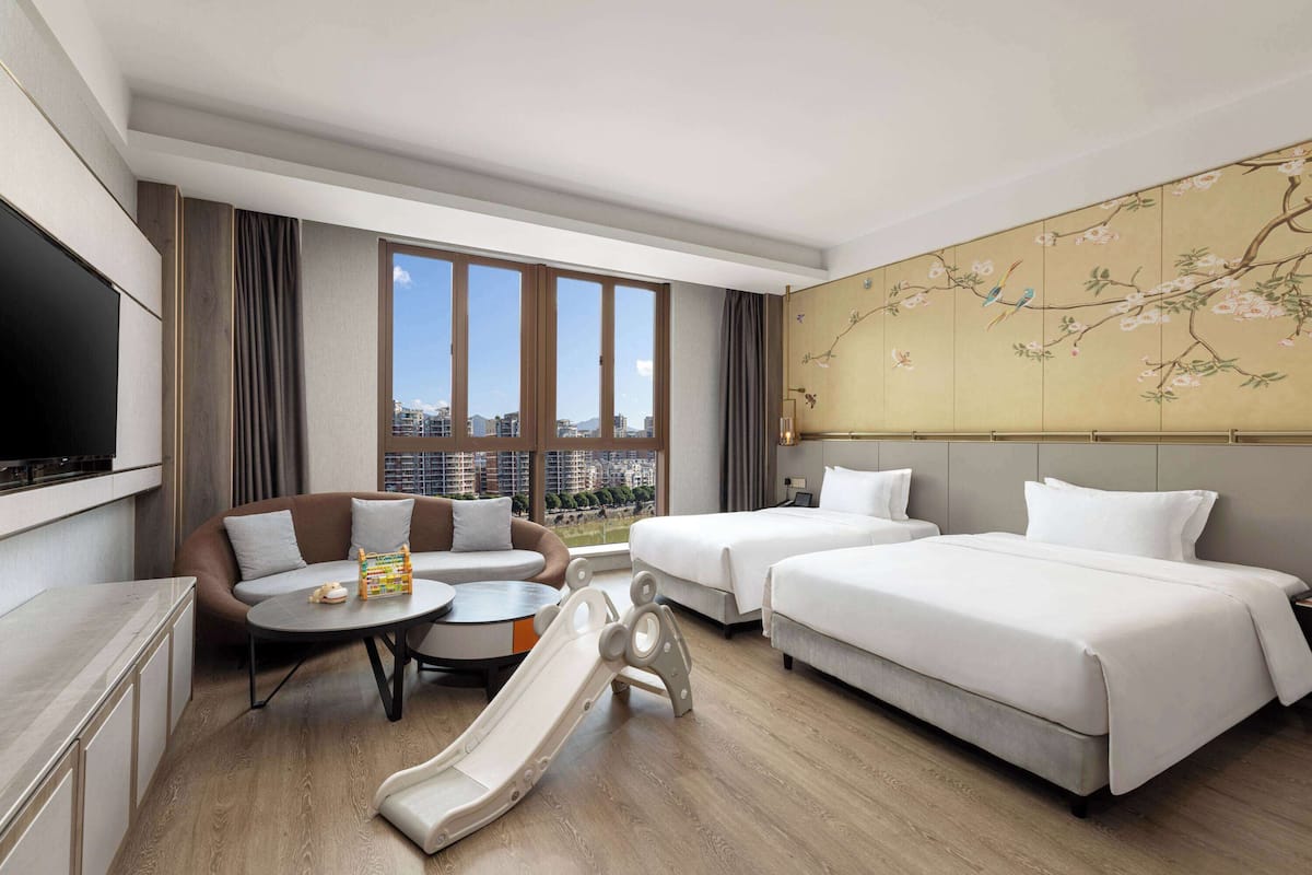 A hotel room with a sofa, two beds, a TV, a dining table, and a view of the city.