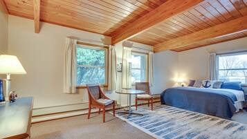 A bedroom with a wooden ceiling, a bed with blue bedding, two chairs, a small table, and a window with curtains.