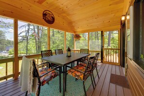 Une terrasse extérieure couverte, dotée d’une table et de chaises, de grandes fenêtres et offrant une vue sur les arbres.