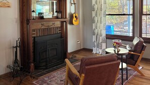 Una sala de estar con chimenea, una guitarra, un sillón de cuero marrón, una mesa redonda de madera y vistas al exterior a través de amplios ventanales.