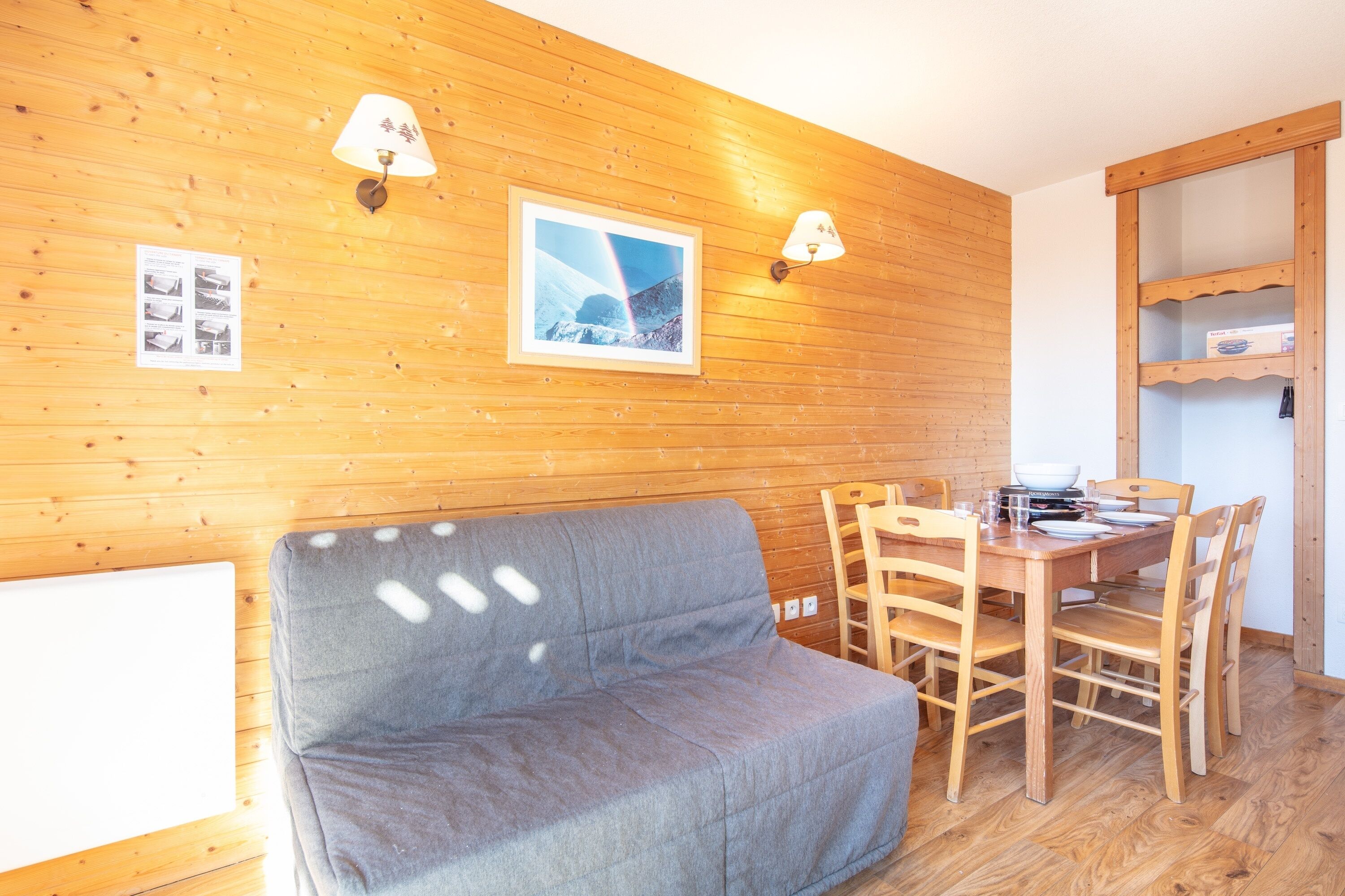 A wooden-paneled room with a grey sofa, wooden dining table with chairs, and wall-mounted lamps.