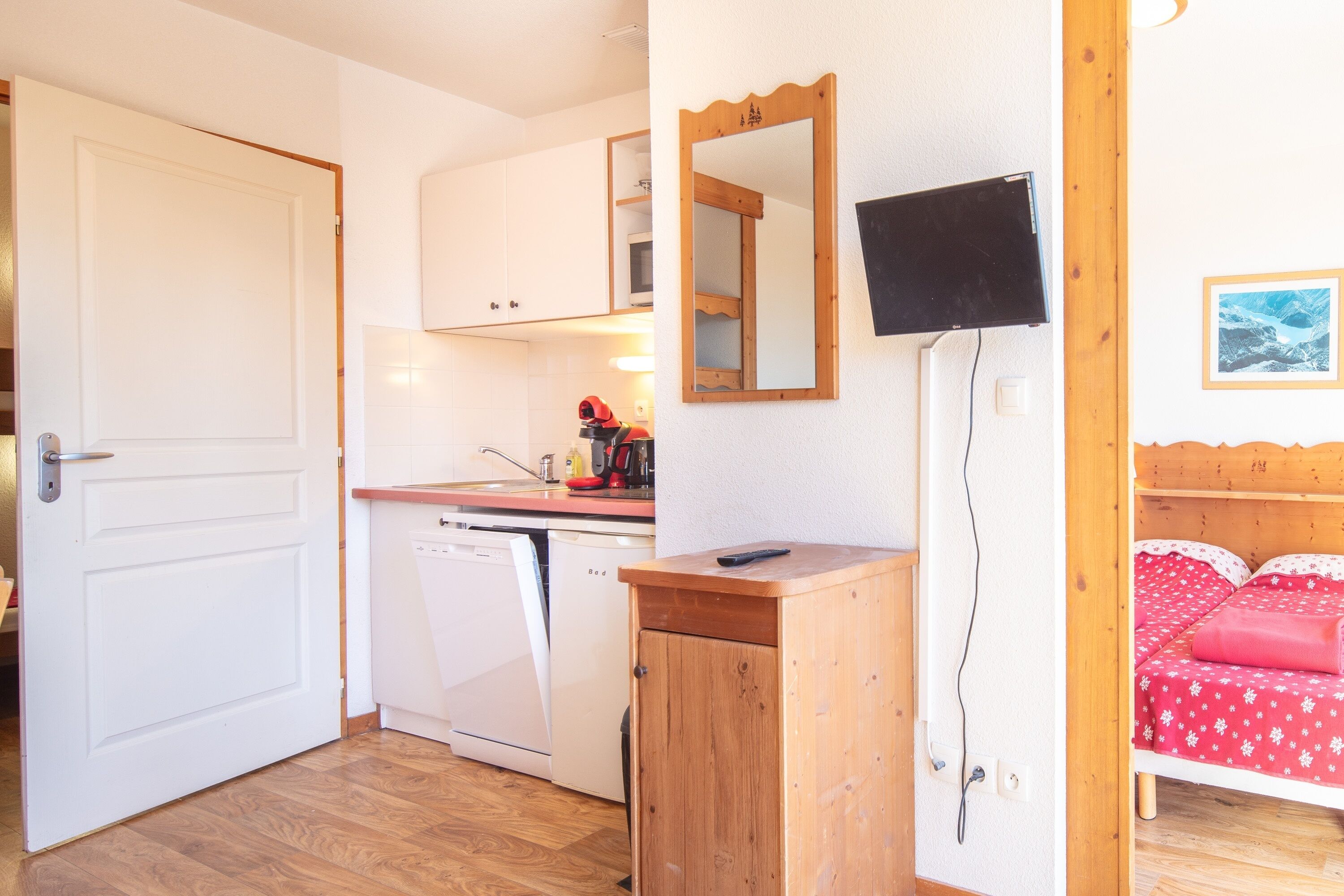 Une pièce compacte avec un bureau en bois, un lit avec une literie rouge, une porte blanche, un coin cuisine équipé d’un lave-vaisselle et un téléviseur fixé au mur.