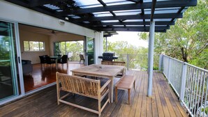 Eine Holzterrasse mit Tisch und Stühlen, einem Grill und einer Glastür, die in einen Innenbereich mit einer Couch und einem Tisch führt.