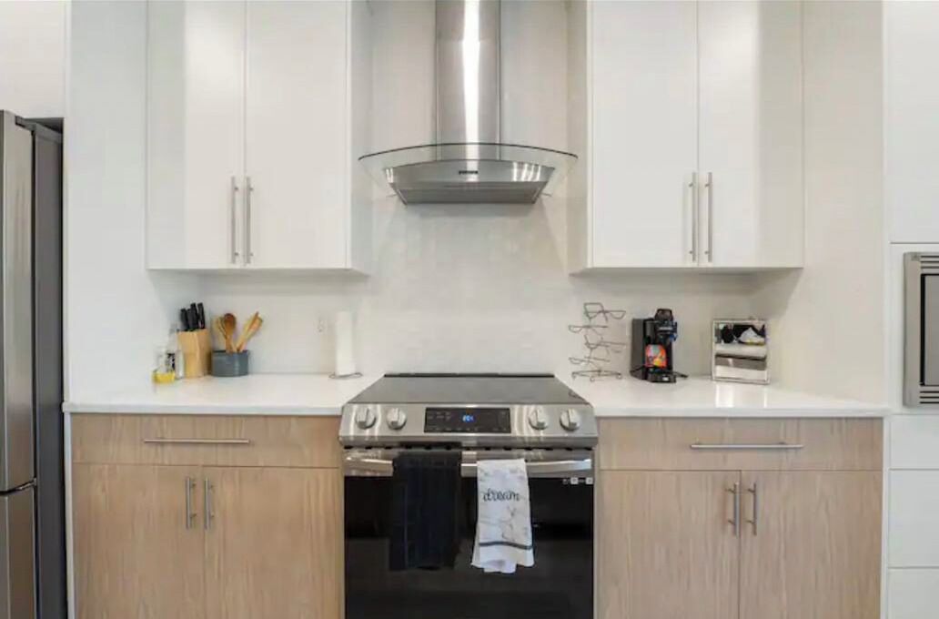 A modern kitchen with white cabinets, a stainless steel range hood, and a built-in oven.