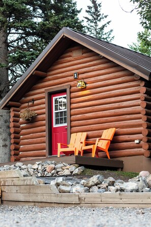 A log cabin with a red door, two orange Adirondack chairs outside, and a sign above the door.