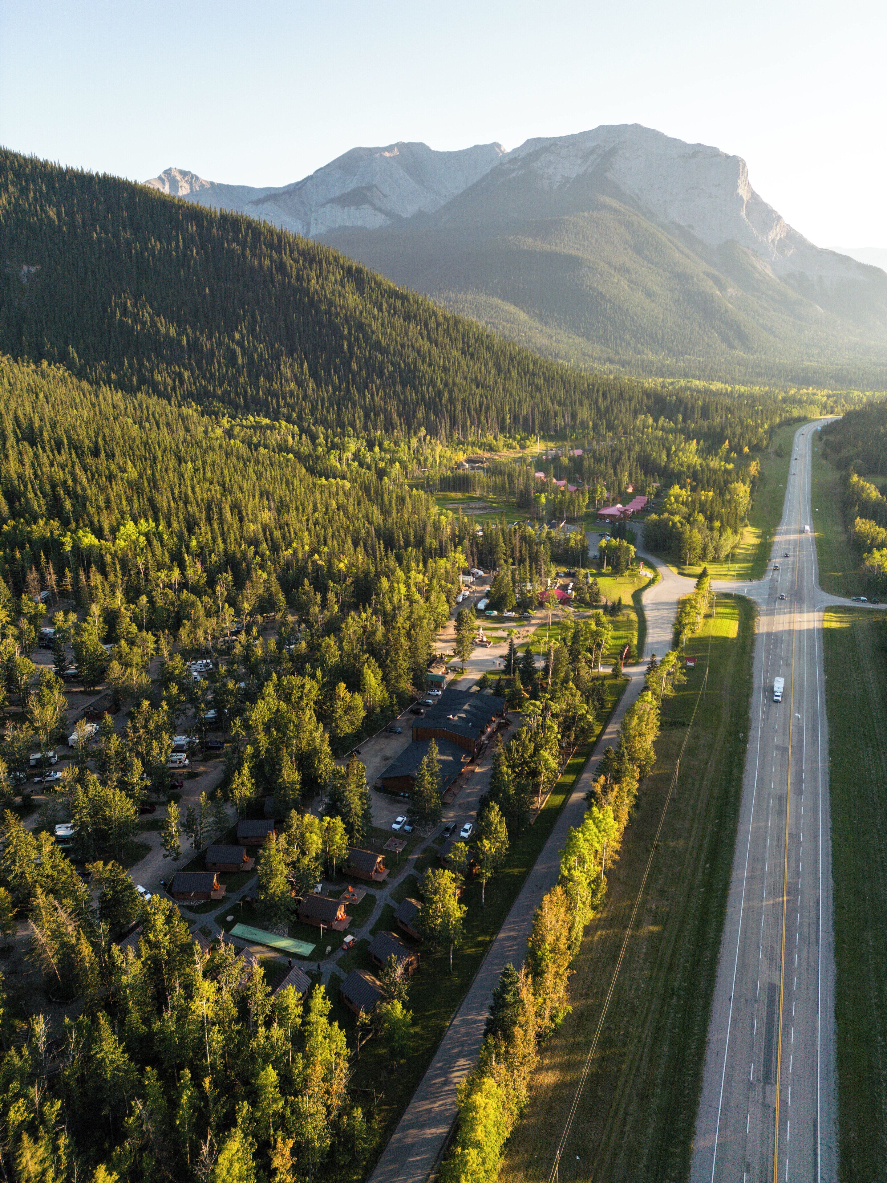 Vista aérea de uma pequena cidade com uma estrada principal, cercada por florestas densas e montanhas ao fundo.