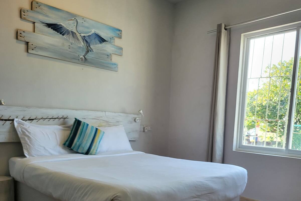 A bedroom with a large bed, a wooden headboard, a painting of birds, a window with curtains, and a view of greenery outside.
