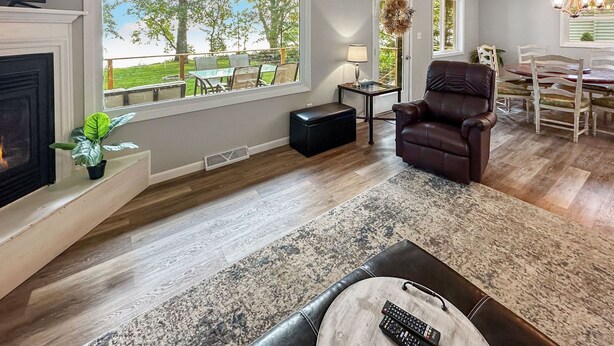 A living room with a fireplace, a leather armchair, a dining area with a table and chairs, and a large window with a view of the outdoors.