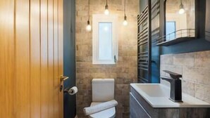 Un baño con puerta de madera, inodoro, lavamanos con espejo e iluminación colgante.
