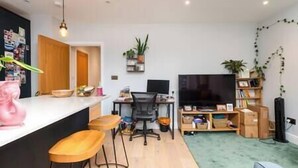 Una cocina moderna con encimera blanca, taburetes de madera, una silla de oficina negra, un televisor y una estantería.