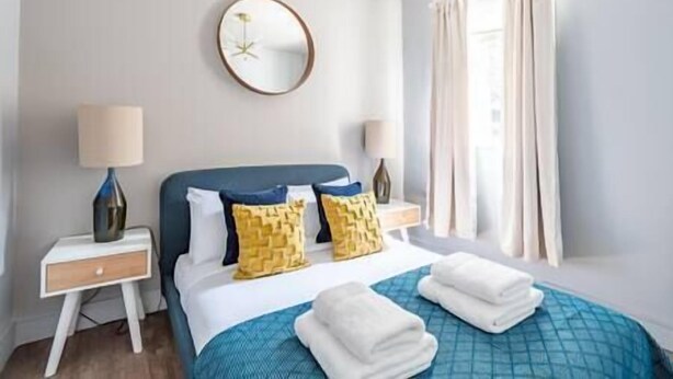 Una habitación de hotel con una cama, dos mesitas de noche, un espejo redondo y un ventanal con cortinas.