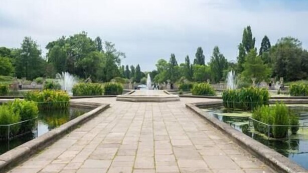 Un sendero pavimentado que conduce a una fuente central en un parque con fuentes de agua y vegetación.