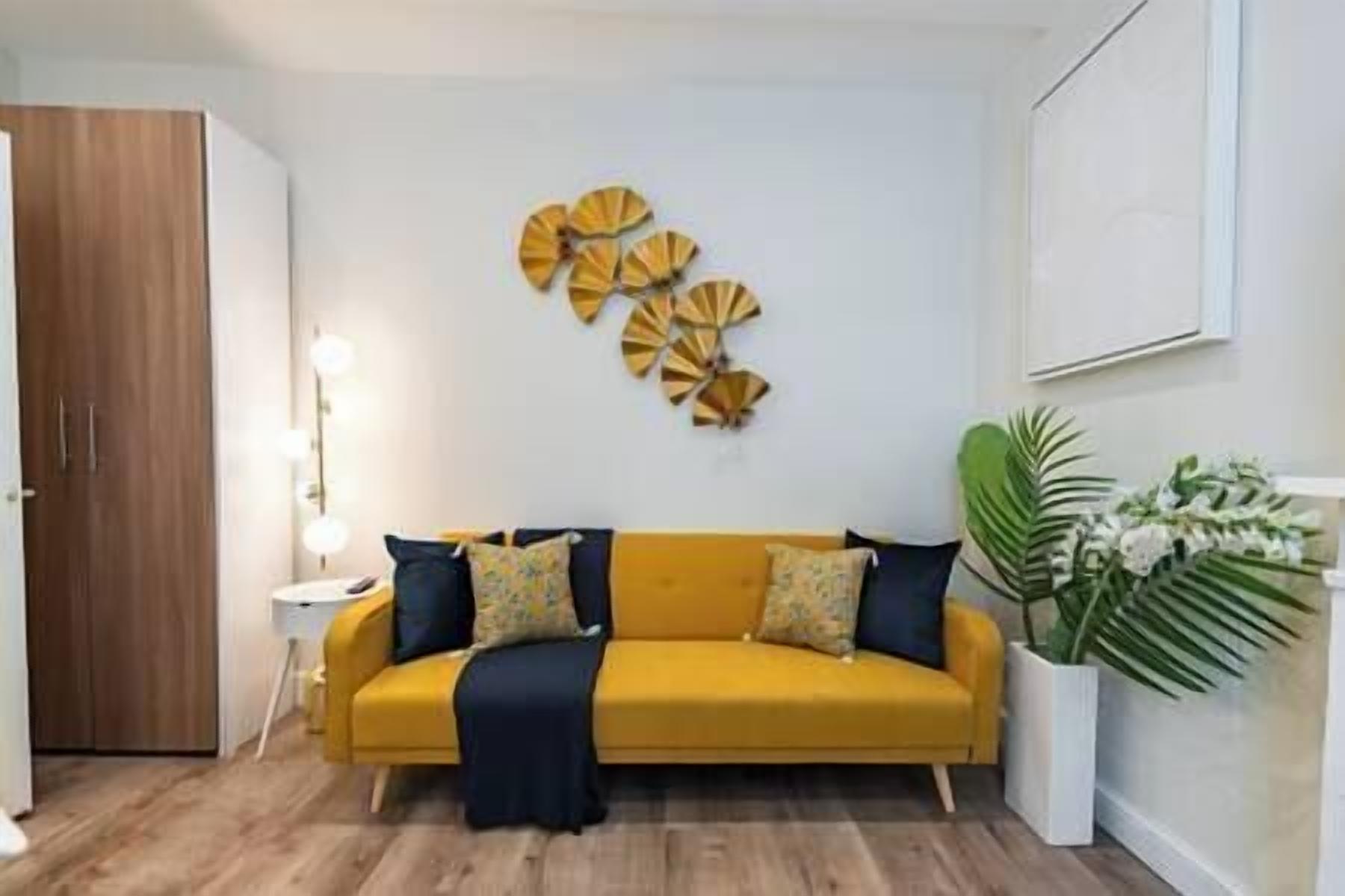A living room with a yellow sofa, dark cushions, a decorative wall piece, a potted plant, and a wooden wardrobe.