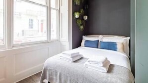 Ein ordentlich bezogenes Bett mit weißen Leinentüchern und einer grauen, strukturierten Bettdecke in einem Raum mit dunkler Wand und einem Fenster mit Blick auf Gebäude.