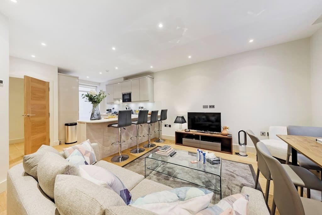 Una sala de estar moderna con un sofá, una mesa de centro de vidrio y un televisor. Hay una zona de cocina con una mesa de comedor y sillas.