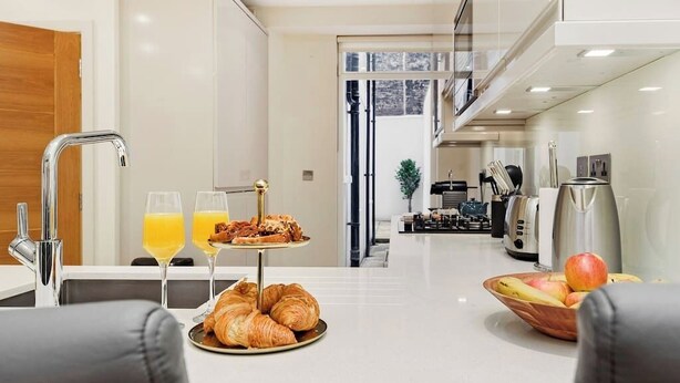 Une cuisine moderne avec un coin petit-déjeuner comprenant des croissants, du jus d’orange et un panier de fruits.