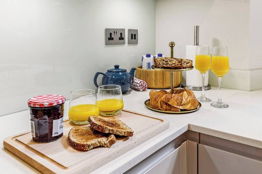 A breakfast setup with toast, jam, orange juice, croissants, and a teapot on a kitchen counter.