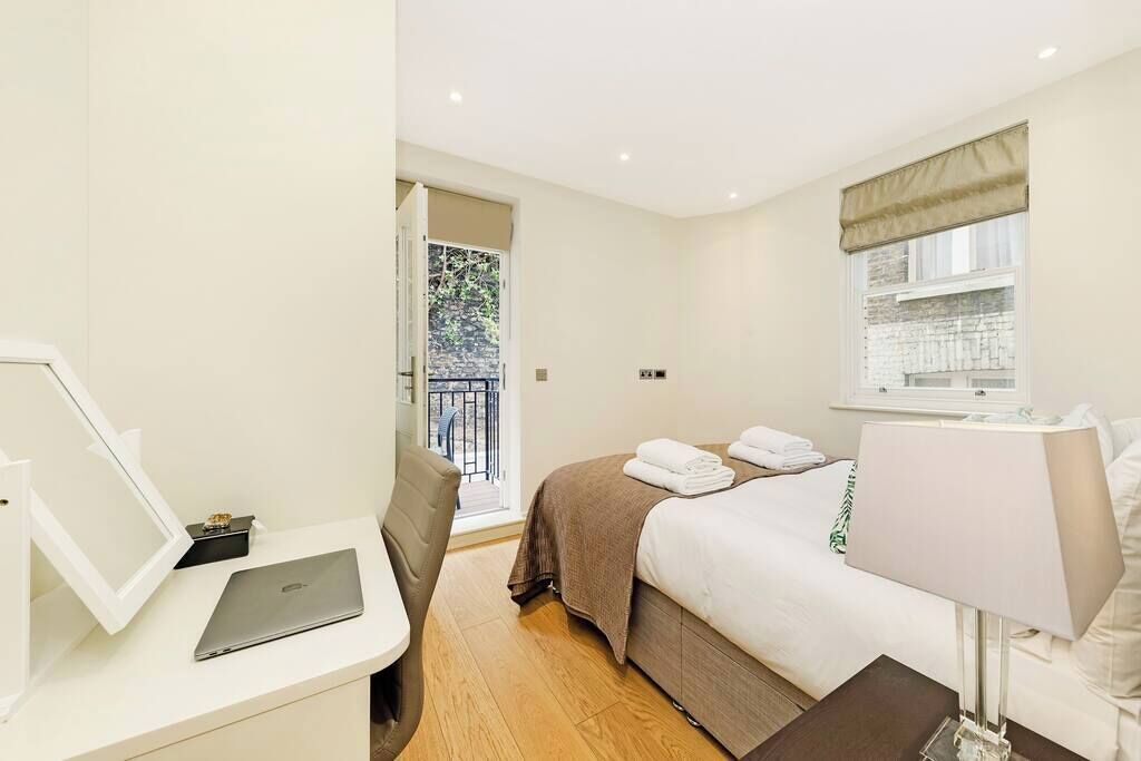 A hotel room with a bed, a desk with a laptop, a chair, a bedside table, and a window with a view of the outside.