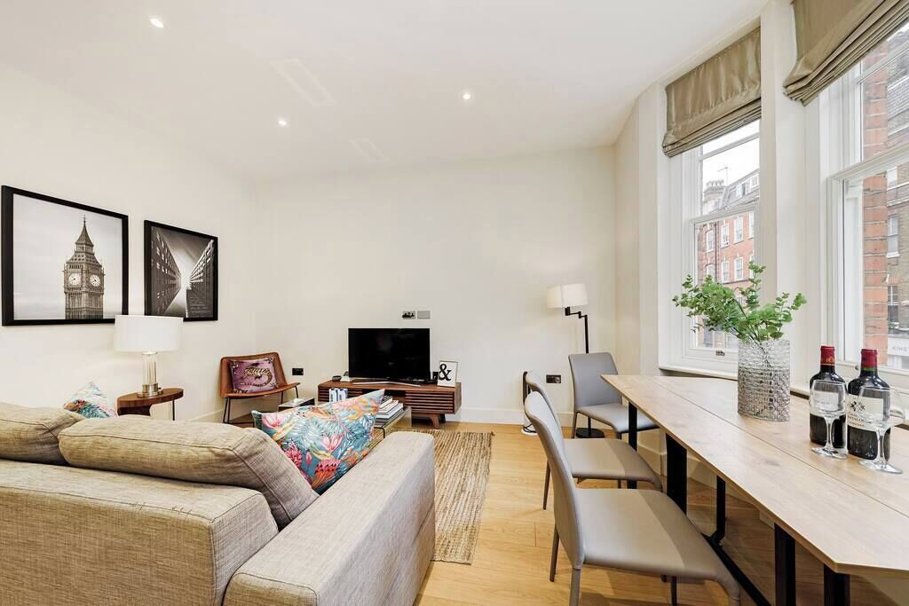 A modern living room with a sofa, a TV, a dining table with chairs, and a large window with a view of buildings.