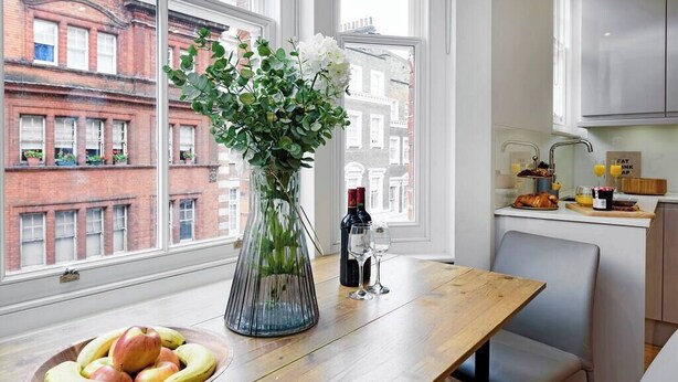 A kitchen with a wooden table, a vase with flowers, a bowl of fruit, and a view of red brick buildings through large windows. - PBC | Modern One Bedroom Apartment in Central London, Fitzrovia GT10 (London)