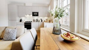 A modern kitchen with white cabinets, a wooden dining table, and a bowl of fruit on the windowsill. - PBC | Modern One Bedroom Apartment in Fitzrovia GT 11 (London)